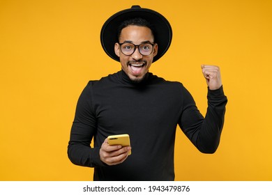 Young smiling happy american african man 20s wear stylish black shirt hat eyeglasses using mobile cell phone do winner gesture clench fist celebrating isolated on yellow background studio portrait.