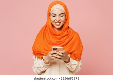 Young smiling happy arabian asian muslim woman wears orange abaya hijab hold in hand use mobile cell phone isolated on plain light pink background. People uae middle eastern islam religious concept - Powered by Shutterstock