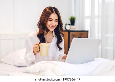 Young smiling happy beautiful asian woman relaxing using laptop computer and drinking coffee in the bedroom at home. Young creative girl working and typing on keyboard.work from home concept
