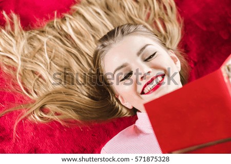 Young smiling happy blonde girl with long hair lying on red background and holding hand made card with lavender in her hands. Valentine's Day, Christmas, Birthday gifts. Red lips