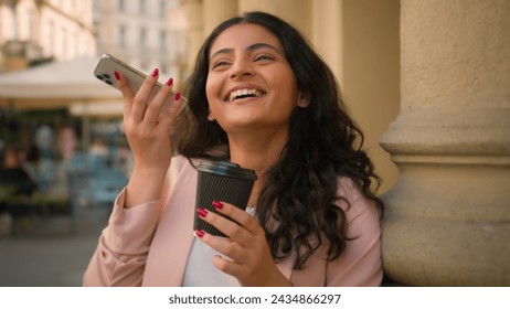 Young smiling happy businesswoman Indian Arabian ethnic woman girl female business worker listening voice message smartphone drink cup coffee receiving audio using cellphone social media outside town - Powered by Shutterstock