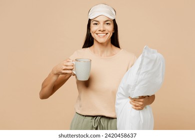 Young smiling happy calm Latin woman wear pyjamas jam sleep eye mask rest relax at home hold pillow cup drink coffee isolated on plain pastel light beige background studio. Good mood night nap concept