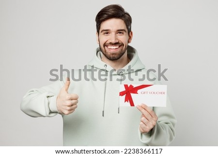 Young smiling happy caucasian man wear mint hoody hold gift certificate coupon voucher card for store show thumb up isolated on plain solid white background studio portrait. People lifestyle concept