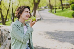 Young Smiling Happy Caucasian Student Woman 20s In Casual Green Jacket Jeans Sitting On Bench In City Spring Park Outdoors Resting Eat Apple Fruit. People Vegeterian Healthy Urban Lifestyle Concept.
