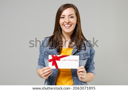 Young smiling happy caucasian woman 20s in casual trendy denim jacket yellow t-shirt holding in hand gift voucher flyer mock up look camera isolated on grey background studio. People lifestyle concept
