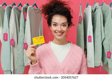 Young Smiling Happy Cheerful Female Costumer Woman Wear Sweater Stand Near Clothes Rack With Tag Sale In Store Showroom Hold In Hand Credit Bank Card Isolated On Plain Pink Background Studio Portrait