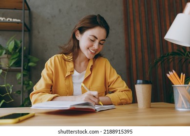 Young smiling happy cheerful fun european successful employee business woman 20s she wearing casual yellow shirt writing in notebook diary sit work at wooden office desk. Achievement career concept