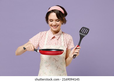 Young smiling happy cheerful housewife housekeeper chef cook baker woman in pink apron holding red frying pan spatula isolated on pastel violet background studio portrait Cooking food process concept.