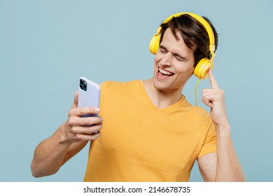 Young Smiling Happy Cheerful Man 20s Wear Yellow T-shirt Headphones Listen To Music Dance Use Mobile Cell Phone Isolated On Plain Pastel Light Blue Background Studio Portrait. People Lifestyle Concept