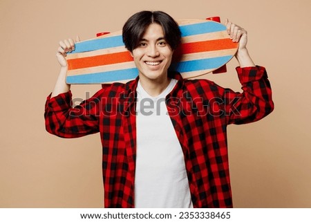 Young smiling happy cheerful man of Asian ethnicity he wear red shirt casual clothes hold skateboard pennyboard look camera isolated on plain pastel light beige background studio. Lifestyle concept