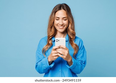 Young smiling happy cheerful woman she wear shirt casual clothes hold in hand use mobile cell phone chat online isolated on plain pastel light blue cyan background studio portrait. Lifestyle concept