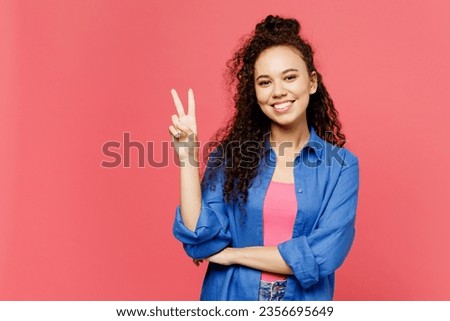 Young smiling happy cheerful woman of African American ethnicity she wear blue shirt casual clothes showing victory sign look camera isolated on plain pastel pink background studio. Lifestyle concept