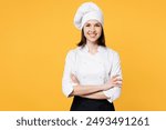 Young smiling happy chef cook baker woman wear white shirt black apron uniform toque chefs hat hold hands crossed folded look camera isolated on plain yellow background studio. Cooking food concept