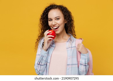 Young Smiling Happy Cool Black Teen Girl Student She Wearing Casual Clothes Backpack Bag Holding Eat Biting Red Apple Isolated On Plain Yellow Color Background. High School University College Concept