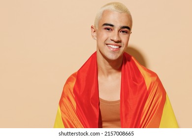 Young smiling happy cool blond latin gay man 20s with make up in beige tank shirt wrapped in rainbow flag looking camera isolated on plain light ocher background studio People lgbt lifestyle concept
