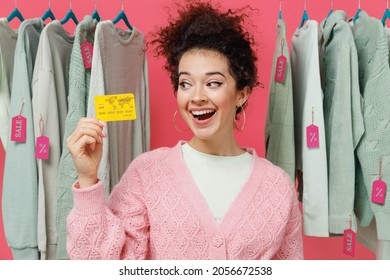 Young Smiling Happy Female Costumer Woman Wear Sweater Stand Near Clothes Rack With Tag Sale In Store Showroom Hold In Hand Look At Credit Bank Card Isolated On Plain Pink Background Studio Portrait