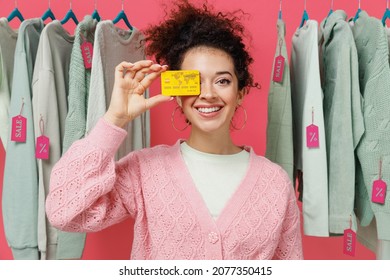 Young Smiling Happy Female Costumer Woman Wear Sweater Stand Near Clothes Rack With Tag Sale In Store Showroom Hold Cover Eye With Credit Bank Card Isolated On Plain Pink Background Studio Portrait