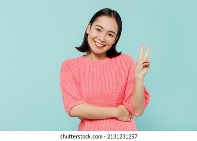 Young smiling happy friendly cool fun woman of Asian ethnicity 20s in pink sweater showing victory sign isolated on pastel plain light blue color background studio portrait. People lifestyle concept