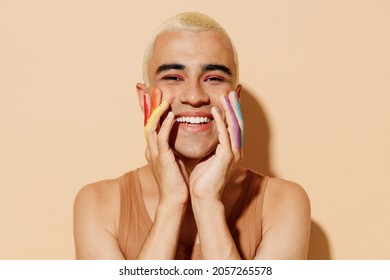 Young smiling happy fun blond latin gay man 20s wearing beige tank shirt posing with make up fingers painted in rainbow flag colors hold face isolated on plain light ocher background studio portrait.