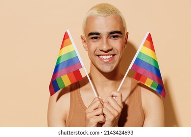 Young Smiling Happy Fun Cheerful Blond Latin Gay Man With Make Up In Beige Tank Shirt Holding Rainbow Flag Isolated On Plain Light Ocher Color Background Studio Portrait People Lgbt Lifestyle Concept