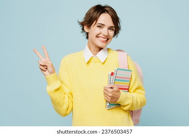 Young smiling happy fun cheerful woman student wear casual clothes yellow sweater backpack bag hold books show victory sign isolated on plain blue background. High school university college concept