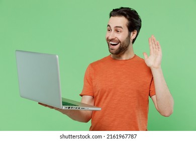 Young Smiling Happy Fun Cool Man 20s In Casual Orange T-shirt Hold Use Work On Laptop Pc Computer Waving Hand Talk By Video Call Isolated On Plain Pastel Light Green Color Background Studio Portrait.