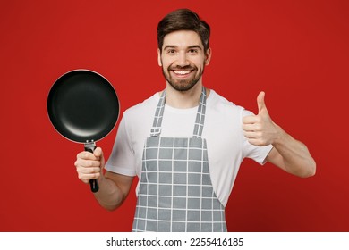 Joven sonriente feliz ama de casa hombre ama de casa chef cocinero hombre panadero vestido con delantal gris sosteniendo en la mano con una sartén de pan show thumb up gesrture aislado en un estudio de fondo rojo plano. Concepto de cocina