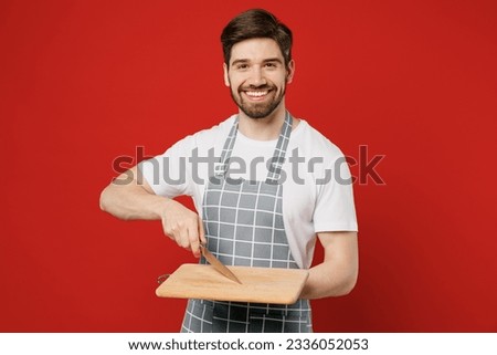 Young smiling happy fun male housewife housekeeper chef cook baker man wear grey apron hold in hand knife empty cutting board look camera isolated on plain red background studio. Cooking food concept