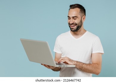 Young Smiling Happy Fun Man 20s Wear Blank Print Design White T-shirt Hold Use Work On Laptop Pc Computer Isolated On Plain Pastel Light Blue Color Background Studio Portrait. People Lifestyle Concept