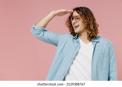 Young Smiling Happy Fun Man 20s With Long Curly Hair In Blue Shirt White T-shirt Glasses Hold Hand At Forehead Look Far Away Distance Isolated On Pastel Plain Pink Color Background Studio Portrait.