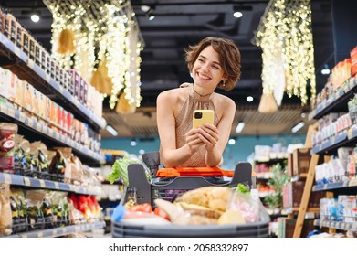 Young Smiling Happy Fun Minded Woman In Casual Clothes Shopping At Supermaket Store With Grocery Cart Using Mobile Cell Phone Look Aside Inside Hypermarket. People Purchasing Gastronomy Food Concept.