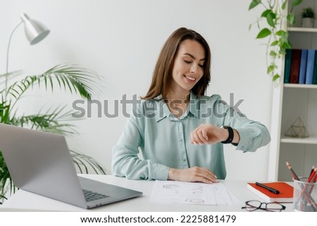 Young smiling happy fun successful employee business woman in blue shirt look at smartwatch time sit work at workplace white desk with laptop pc computer at office indoors. Achievement career concept