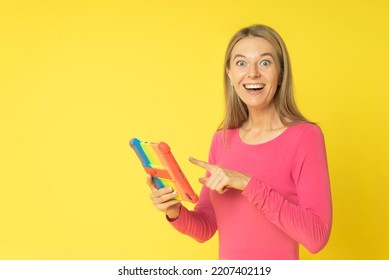 Young Smiling Happy Fun Woman 30s Wearing Pink Dress Holds Tablet Computer In Colored Cover Pointing Index Finger Isolated On Yellow Color Background Studio Portrait.