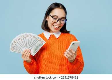Young smiling happy fun woman of Asian ethnicity wear orange sweater glasses use mobile cell phone hold fan of cash money in dollar banknotes isolated on plain pastel light blue cyan background studio - Shutterstock ID 2251578411