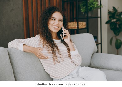 Young smiling happy fun woman wear casual clothes sits on grey sofa couch talk speak on mobile cell phone stay at home hotel flat rest relax spend free spare time in living room indoor. Lounge concept - Powered by Shutterstock