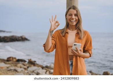 Young smiling happy fun woman she wears orange shirt casual clothes hold in hand use mobile cell phone show ok okay walk on sea ocean sand shore beach outdoor seaside in summer day. Lifestyle concept - Powered by Shutterstock