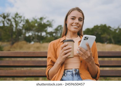 Young smiling happy fun woman wears orange shirt casual clothes use mobile cell phone drink coffee sit on bench walk rest relax in spring green city park outdoors on nature. Urban lifestyle concept - Powered by Shutterstock