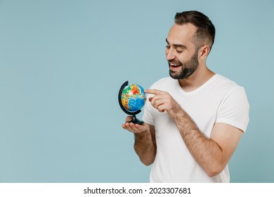Young smiling happy geography teacher man 20s wear casual white t-shirt hold in hands touch hoose country spin Earth world globe isolated on plain pastel light blue color background studio portrait.