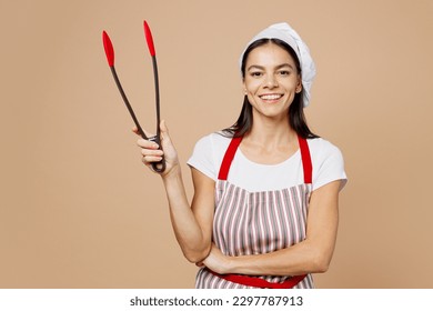 Young smiling happy housewife housekeeper chef baker latin woman wear striped apron toque hat hold in hand barbecue tongs look camera isolated on plain pastel light beige background. Cook food concept - Powered by Shutterstock