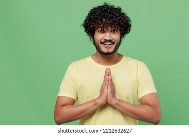 Young Smiling Happy Indian Man 20s In Basic Yellow T-shirt Hold Hands Folded In Prayer Gesture, Begging About Something Isolated On Plain Pastel Light Green Background Studio People Lifestyle Concept