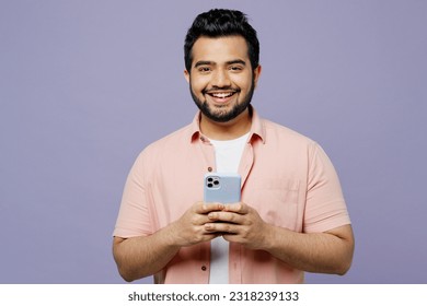 Young smiling happy Indian man he wear pink shirt white t-shirt casual clothes hold in hand use mobile cell phone look camera isolated on plain pastel light purple background studio. Lifestyle concept - Shutterstock ID 2318239133