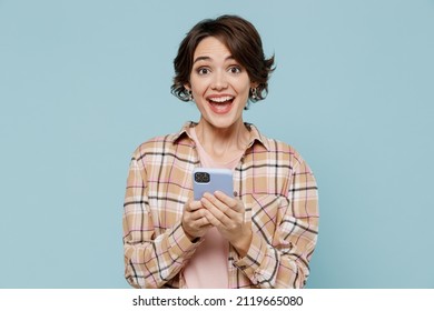 Young Smiling Happy Joyful Fun Woman 20s Wearing Casual Brown Shirt Hold In Hand Use Mobile Cell Phone Isolated On Pastel Plain Light Blue Color Background Studio Portrait. People Lifestyle Concept