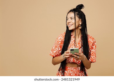 Young smiling happy Latin woman she wears orange casual clothes hold in hand use mobile cell phone look aside on area isolated on plain pastel light beige background studio portrait. Lifestyle concept - Powered by Shutterstock