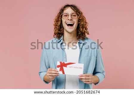 Young smiling happy man 20s with long curly hair in blue shirt white t-shirt glasses hold gift certificate coupon voucher card for store isolated on pastel plain pink color background studio portrait.