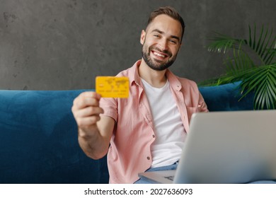 Young Smiling Happy Man In Casual Clothes Using Work On Laptop Pc Computer Give Credit Bank Card Shopping Online Order Delivery Booking Tour Sitting On Blue Sofa At Home Flat Indoors Rest On Weekends