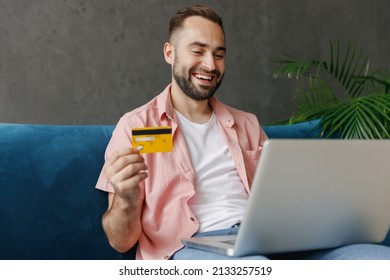 Young Smiling Happy Man In Casual Clothes Using Work On Laptop Pc Computer Credit Bank Card Shopping Online Order Delivery Booking Tour Sitting On Blue Sofa At Home Flat Indoors Rest Relax On Weekends