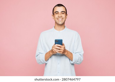 Young smiling happy middle eastern man he wearing casual blue cotton shirt holding in hand use mobile cell phone isolated on plain pastel light pink wall background studio portrait. Lifestyle concept