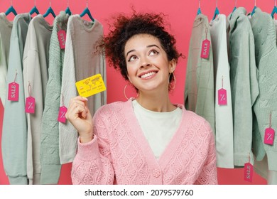 Young Smiling Happy Minded Female Costumer Woman Wear Sweater Stand Near Clothes Rack With Tag Sale In Store Showroom Hold In Hand Credit Bank Card Isolated On Plain Pink Background Studio Portrait