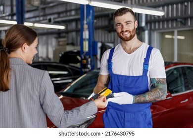 Young Smiling Happy Professional Car Mechanic Man In Blue Overalls Gloves Hold Payment Terminal Fow Paying With Credit Card By Female Owner Driver Woman Work In Vehicle Repair Shop Workshop Indoors.