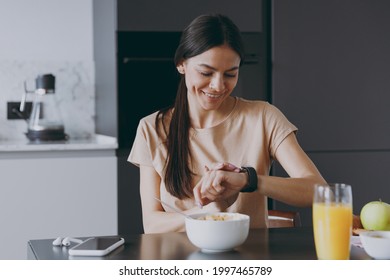 Young Smiling Happy Punctual Calm Housewife Woman 20s Wear Beige T-shirt Eat Breakfast In Morning Look At Smart Watch Check Time Cooking Food In Light Kitchen At Home Healthy Diet Lifestyle Concept